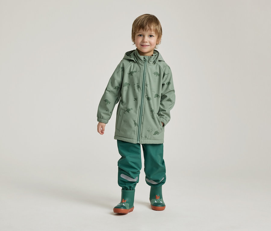Ein Junge steht in grüner Dino-Regenjacke, grüner Hose und Gummistiefeln vor einem hellen Hintergrund.
