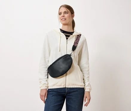 Eine Frau trägt eine schwarze Gürteltasche mit einem Trageriemen mit Leopardenmuster über einer cremefarbenen Jacke und Jeans.