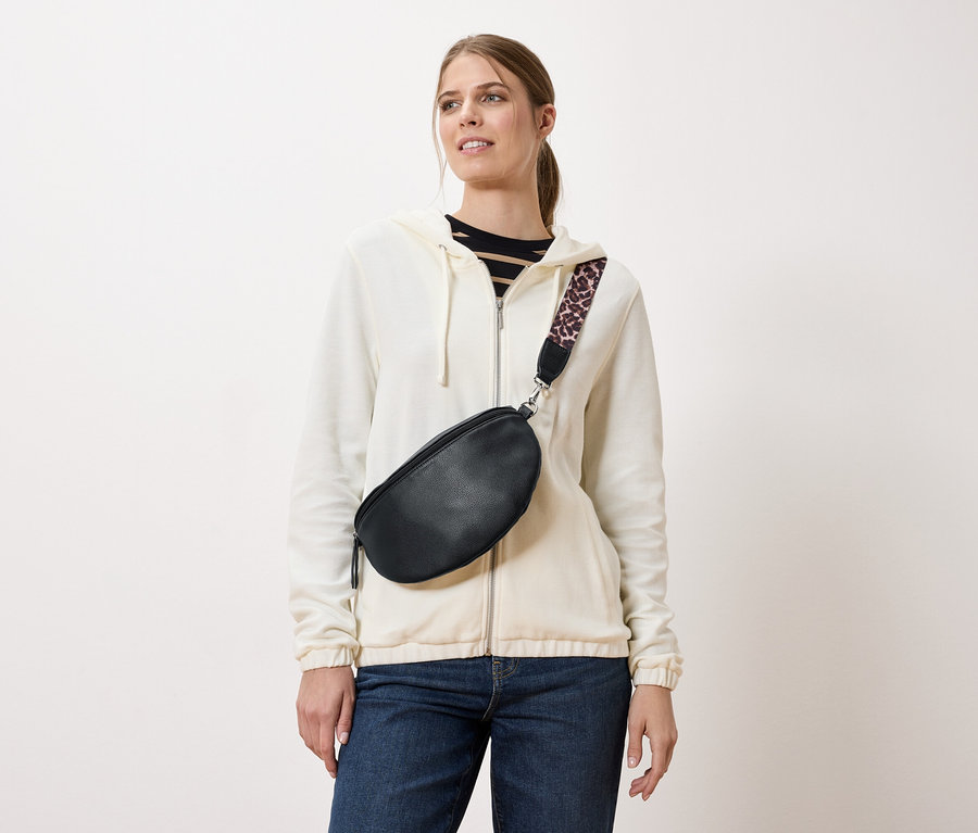 Eine Frau trägt eine schwarze Gürteltasche mit einem Trageriemen mit Leopardenmuster über einer cremefarbenen Jacke und Jeans.