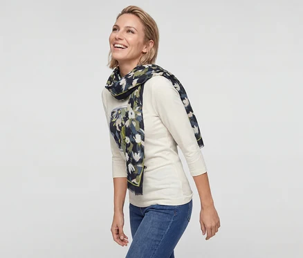 Lächelnde Frau mit kurzen blonden Haaren trägt ein cremefarbenes Langarmshirt, blaue Jeans und einen gemusterten Schal.