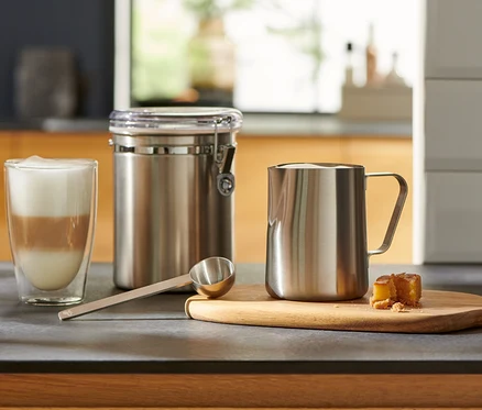 Auf der Küchentheke stehen ein Glas mit Latte Macchiato, ein silbernes Kaffeelot, ein Behälter und ein Milchkännchen.