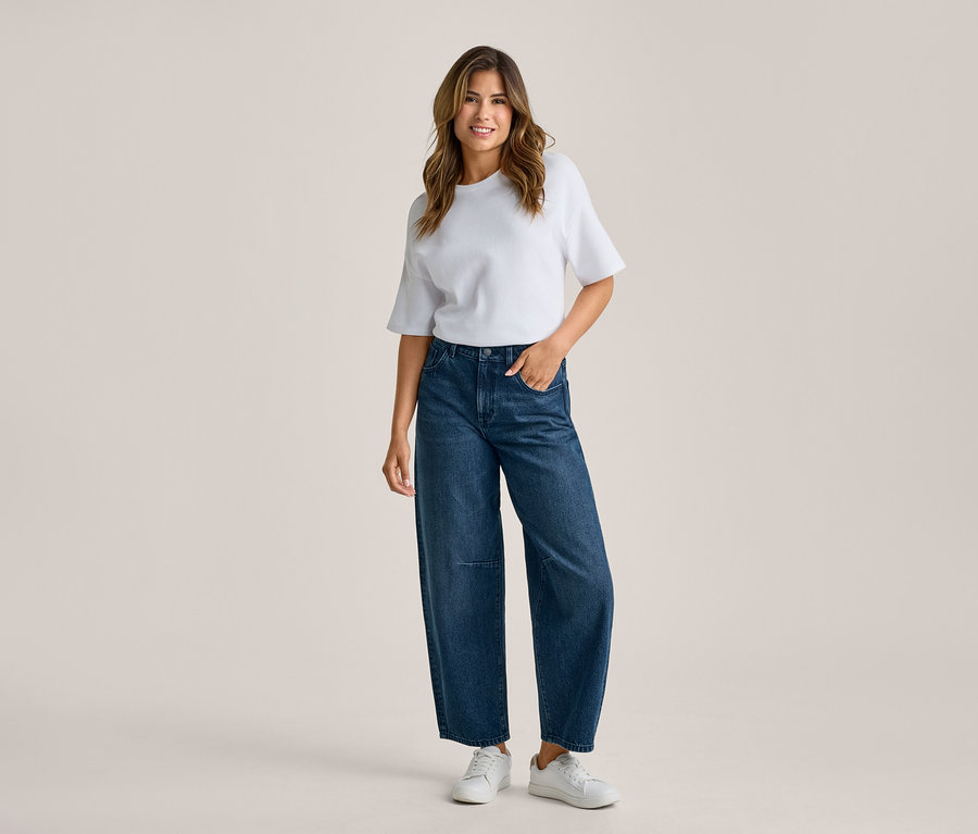 Frau steht lächelnd in weißem T-Shirt, dunkelblauer weiter Jeans und weißen Sneakers, eine Hand in der Tasche.