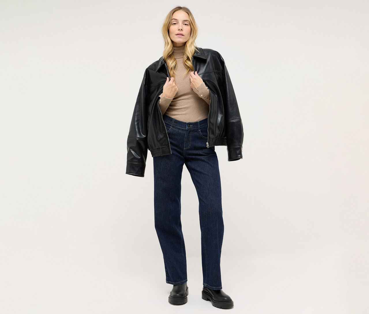 Blonde Frau steht, trägt einen beigen Rollkragenpullover, dunkelblaue Jeans und schwarze Stiefel, eine schwarze Lederjacke über die Schultern gelegt.