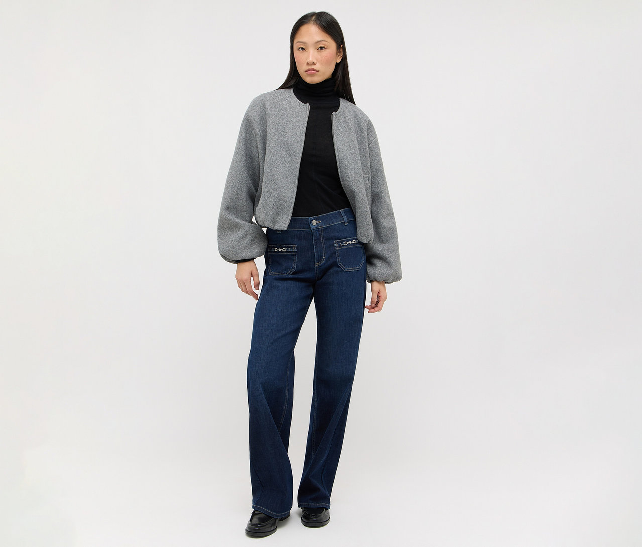 Frau trägt einen schwarzen Rollkragenpullover, eine graue, kurze Jacke mit Puffärmeln, dunkelblaue weite Jeans mit Ziertaschen und schwarze flache Schuhe.