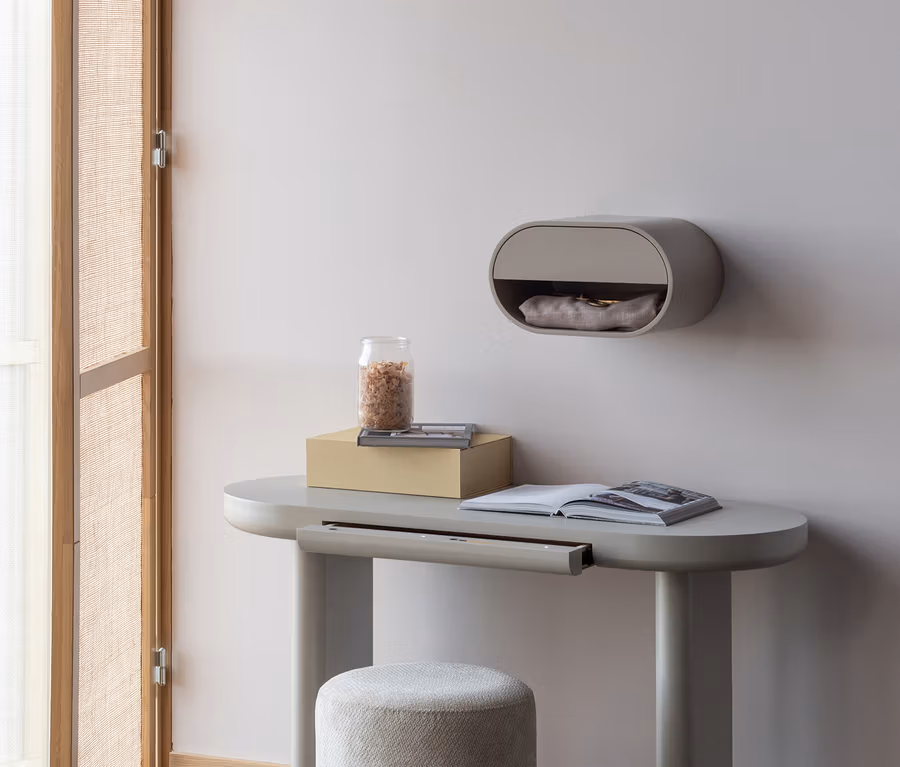 Ein hellgrauer, ovaler Schreibtisch mit einem passenden Hocker steht vor einer hellen Wand. Auf dem Schreibtisch liegen ein offenes Buch, eine gelbe Box und ein Glasgefäß. Darüber hängt ein ovales Wandregal mit einem Fach.