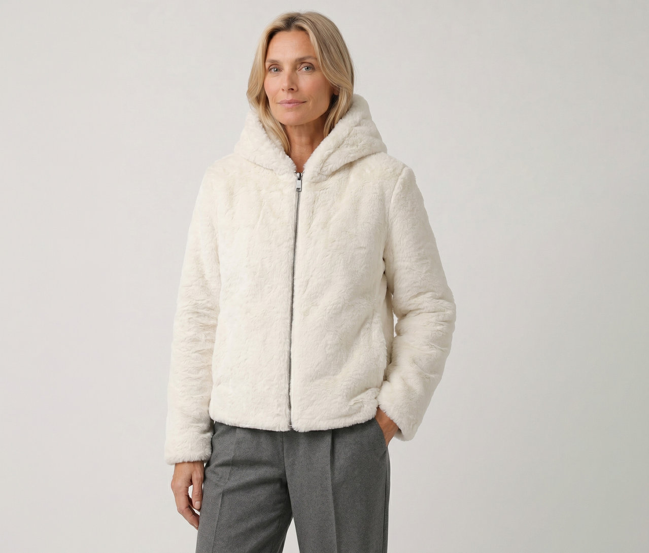 Eine blonde Frau trägt eine cremefarbene, flauschige Kapuzenjacke und graue Hosen, ihre rechte Hand steckt in der Tasche.