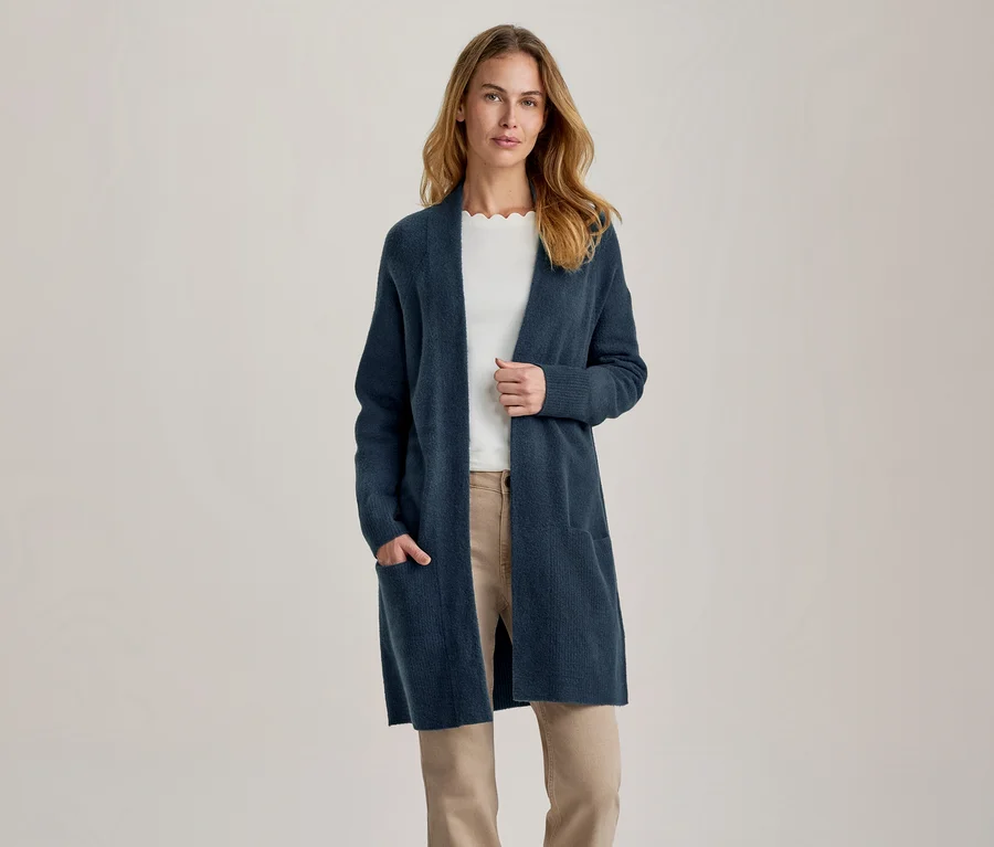 Frau trägt eine lange, dunkelblaue offene Strickjacke über einem weißen Oberteil und beiger Hose, eine Hand in der Tasche.