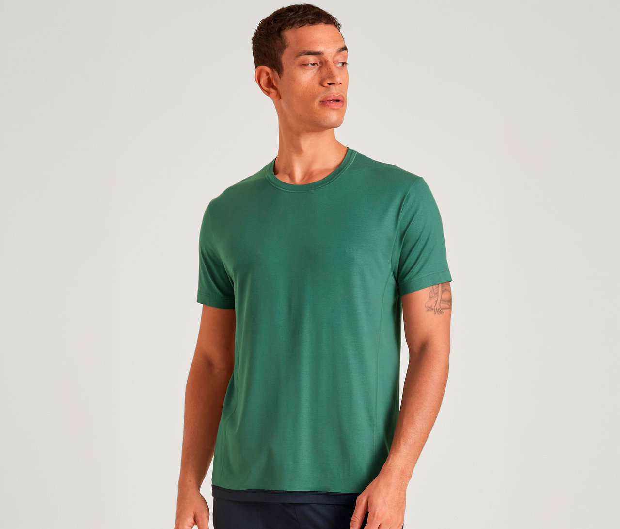Ein Mann mit kurzen dunklen Haaren und einem Tattoo am rechten Arm trägt ein schlichtes grünes Kurzarm-T-Shirt und eine dunkle Hose, blickt nach rechts vor weißem Hintergrund.