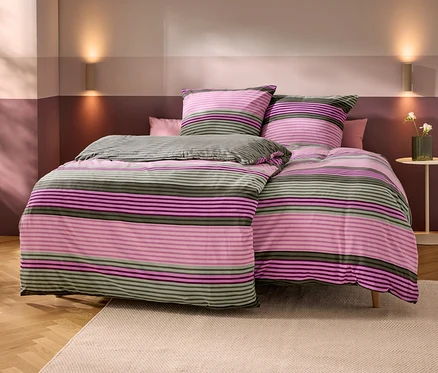Bett mit gestreifter Bettwäsche in Rosa, Lila und Olivgrün. Zwei passende Kissen und ein kleines rosa Kissen liegen darauf. Die Wände sind zweifarbig in Hell- und Dunkelrosa/Violett, mit zwei Wandleuchten. Rechts steht ein Beistelltisch mit einer Vase weißer Blumen. Holzboden mit beigem Teppich.