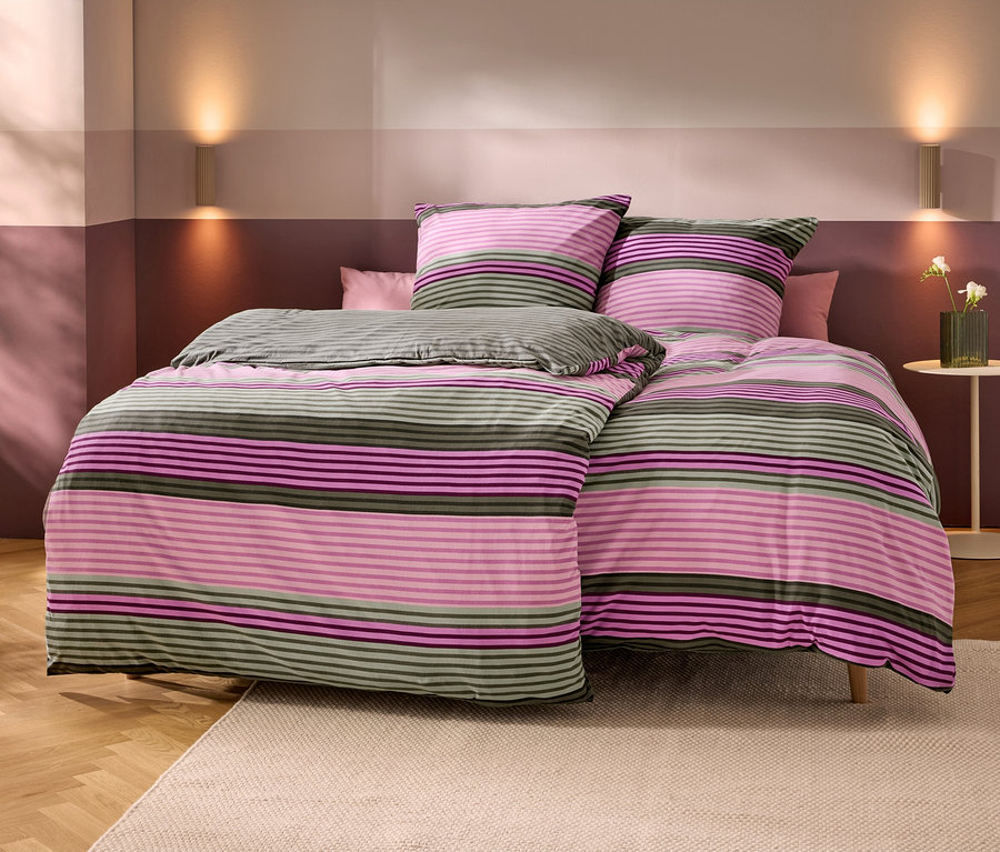 Bett mit gestreifter Bettwäsche in Rosa, Lila und Olivgrün. Zwei passende Kissen und ein kleines rosa Kissen liegen darauf. Die Wände sind zweifarbig in Hell- und Dunkelrosa/Violett, mit zwei Wandleuchten. Rechts steht ein Beistelltisch mit einer Vase weißer Blumen. Holzboden mit beigem Teppich.