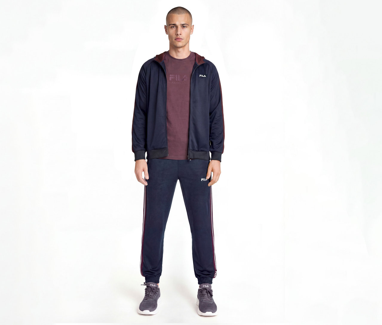 Mann trägt einen marineblauen Fila Trainingsanzug mit bordeauxroten Seitenstreifen, ein bordeauxrotes T-Shirt und graue Turnschuhe, stehend vor weißem Hintergrund.