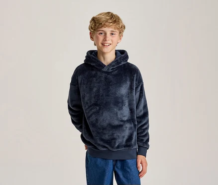 Lächelnder junger Junge mit blonden Locken in einem dunkelblauen flauschigen Hoodie und blauen Jeans, Hände hinter dem Rücken.