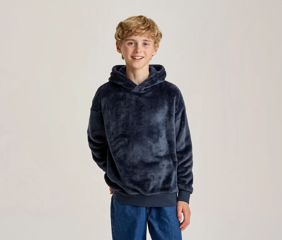 Lächelnder junger Junge mit blonden Locken in einem dunkelblauen flauschigen Hoodie und blauen Jeans, Hände hinter dem Rücken.