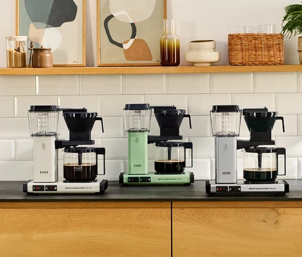 Drei Filterkaffeemaschinen Moccamaster KBG Select, eine weiße, eine mintgrüne und eine silberne, stehen auf einer Küchenarbeitsplatte, jede mit einer Glaskanne Kaffee. Darüber befindet sich ein Holzregal mit Dekorationsartikeln.