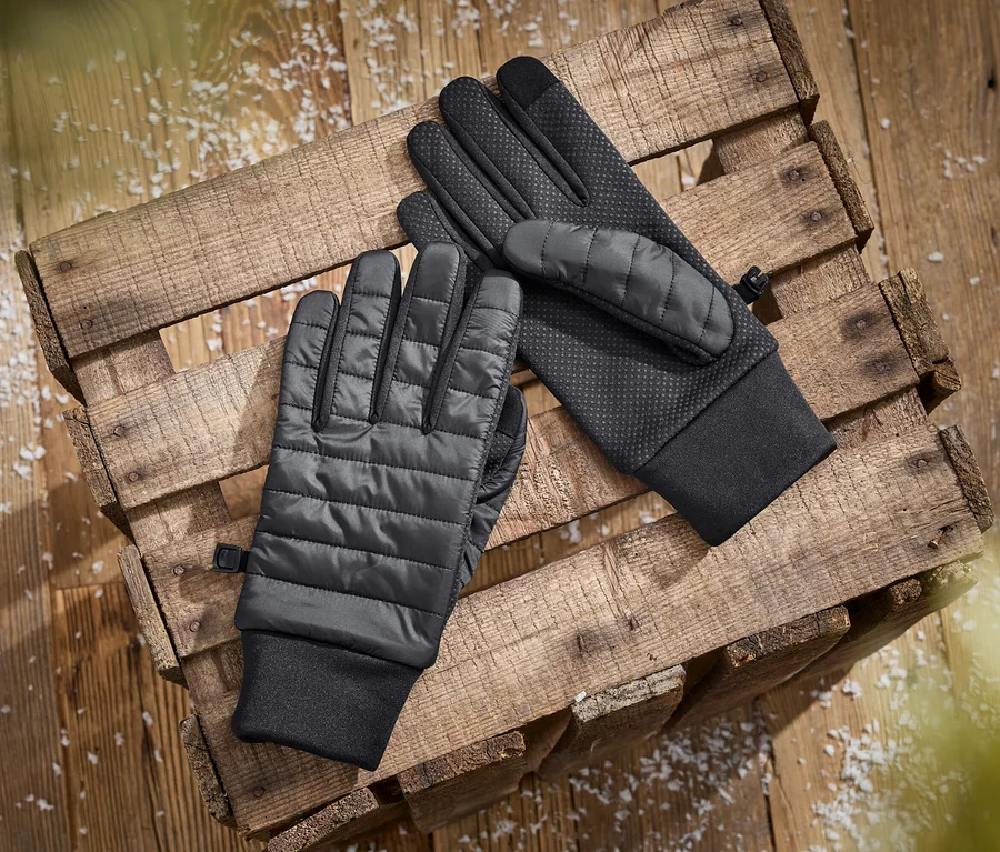 Zwei schwarze Handschuhe, einer mit gestepptem Handrücken und der andere mit texturierter Handfläche, liegen auf einer Holzkiste mit verstreuten weißen Flocken.