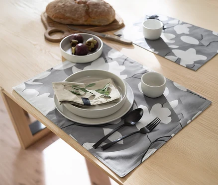 Holztisch mit zwei grauen, floral gemusterten SCHÖNER WOHNEN Tischsets. Ein Set ist mit Tellern, Besteck und Tasse gedeckt, im Hintergrund Brot und Früchte.