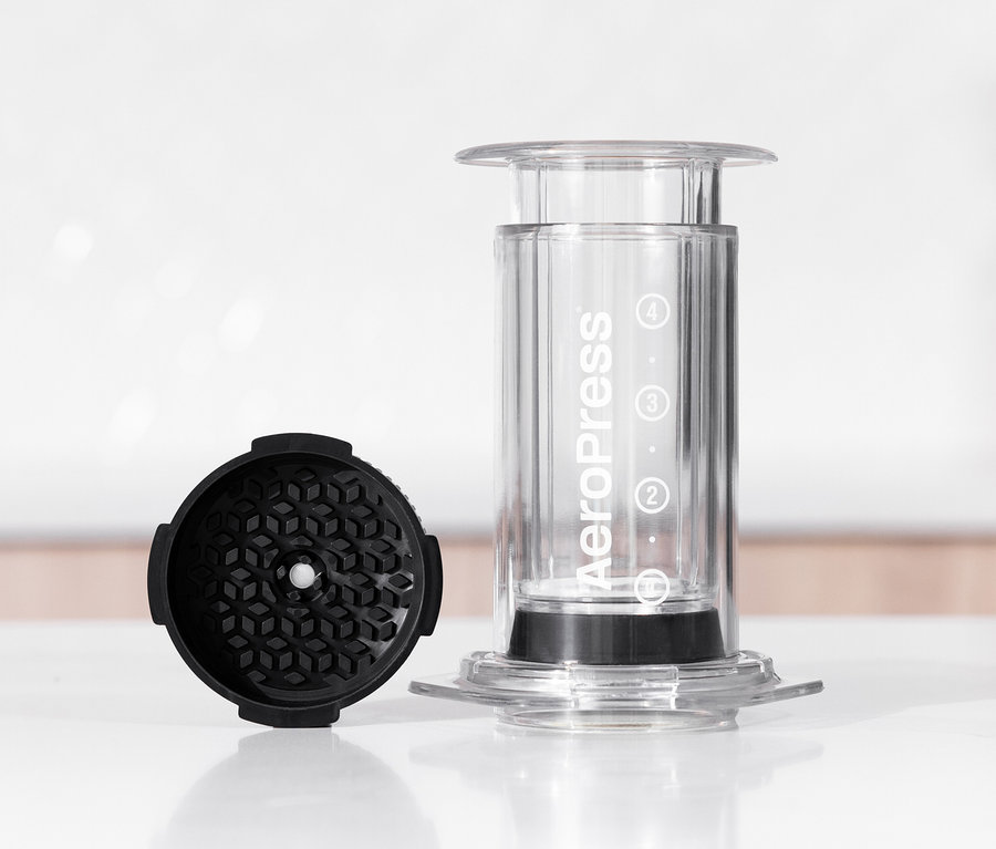 Ein transparenter AeroPress Coffee Maker »Clear« steht neben einem schwarzen Filteraufsatz auf einem Tisch.