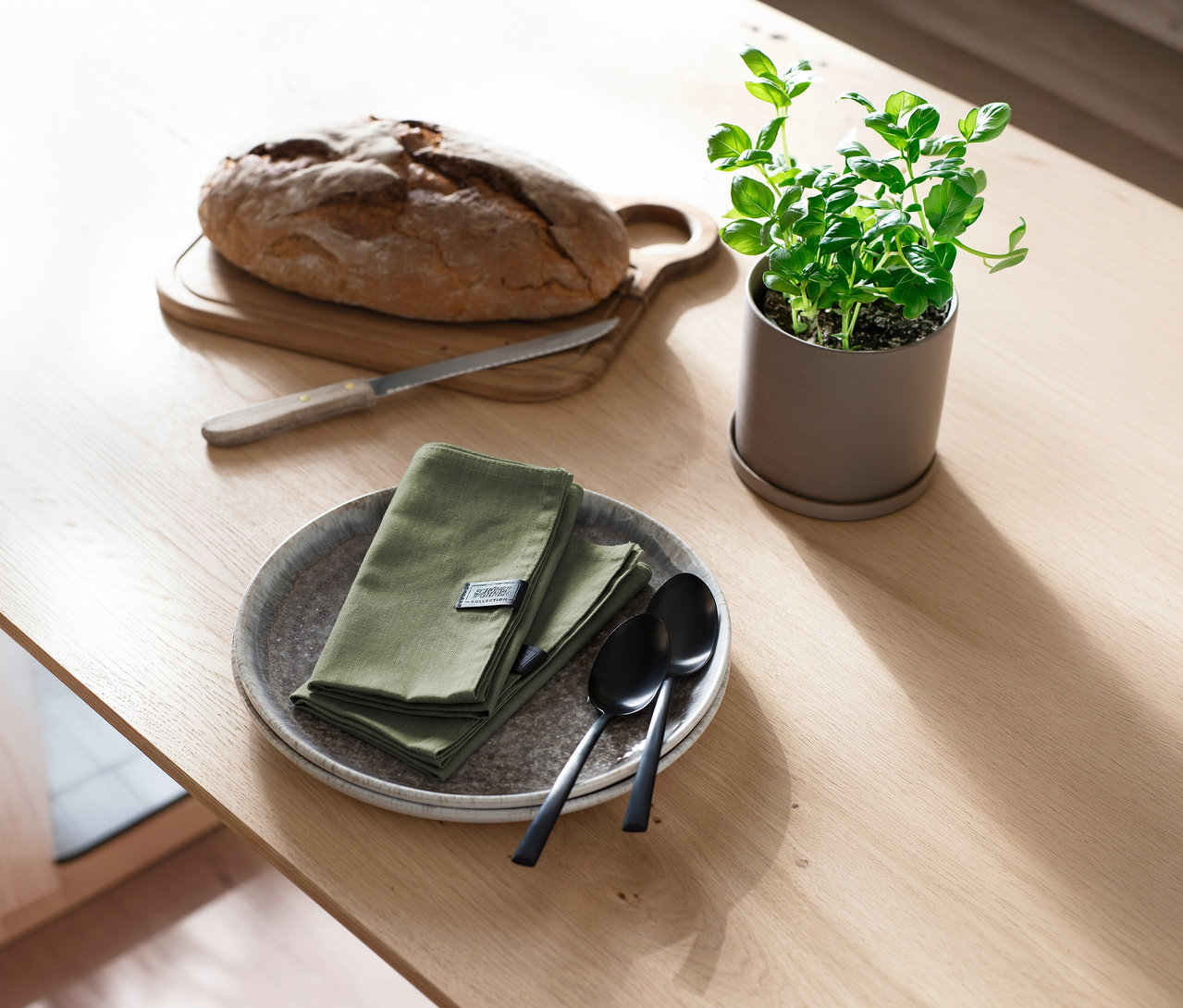 Ein Laib Brot auf einem Holzbrett mit Brotmesser, daneben graue Teller mit gefalteten khakifarbenen Servietten der SCHÖNER WOHNEN-Kollektion und schwarzen Löffeln, sowie eine Topfpflanze auf einem Holztisch.