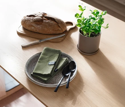 Ein Laib Brot auf einem Holzbrett mit Brotmesser, daneben graue Teller mit gefalteten khakifarbenen Servietten der SCHÖNER WOHNEN-Kollektion und schwarzen Löffeln, sowie eine Topfpflanze auf einem Holztisch.