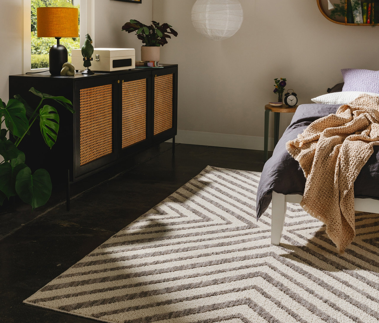 Grau-weißer benuta Teppich »Junis Chevron« mit Zickzackmuster in einem Schlafzimmer, neben einem Bett mit grauer Bettwäsche und einem schwarzen Sideboard.