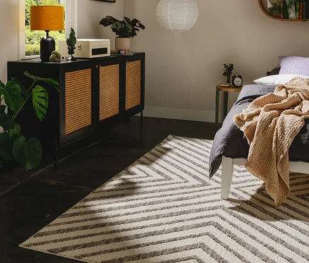 Grau-weißer benuta Teppich »Junis Chevron« mit Zickzackmuster in einem Schlafzimmer, neben einem Bett mit grauer Bettwäsche und einem schwarzen Sideboard.