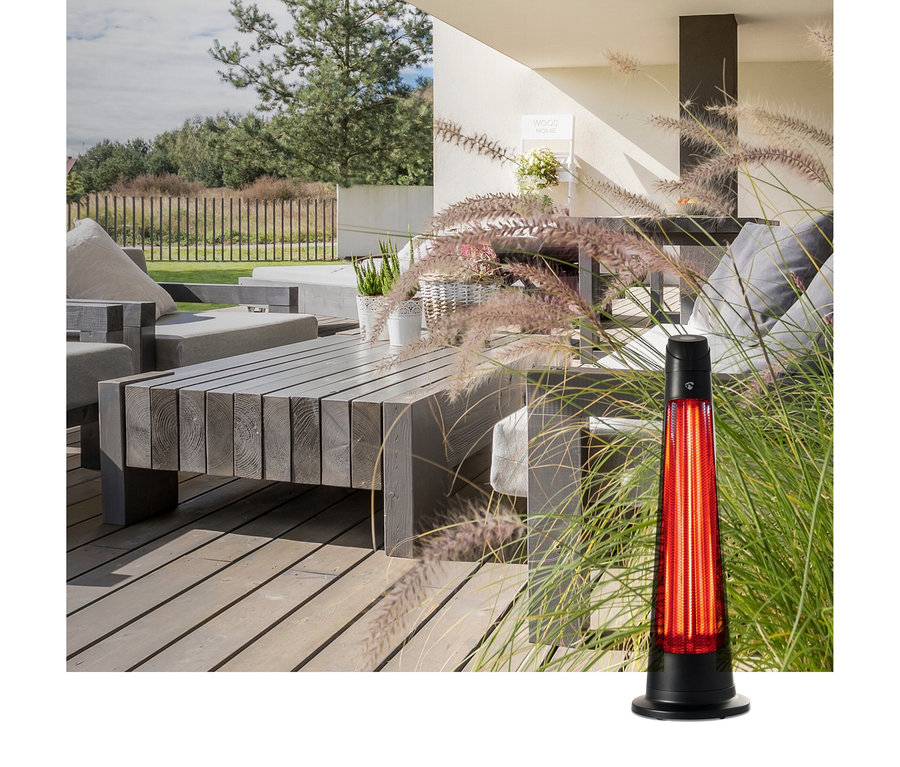 Ein schwarzer Terrassenheizer mit rot leuchtendem Element steht im hohen Gras auf einer Holzterrasse mit Lounge-Möbeln.