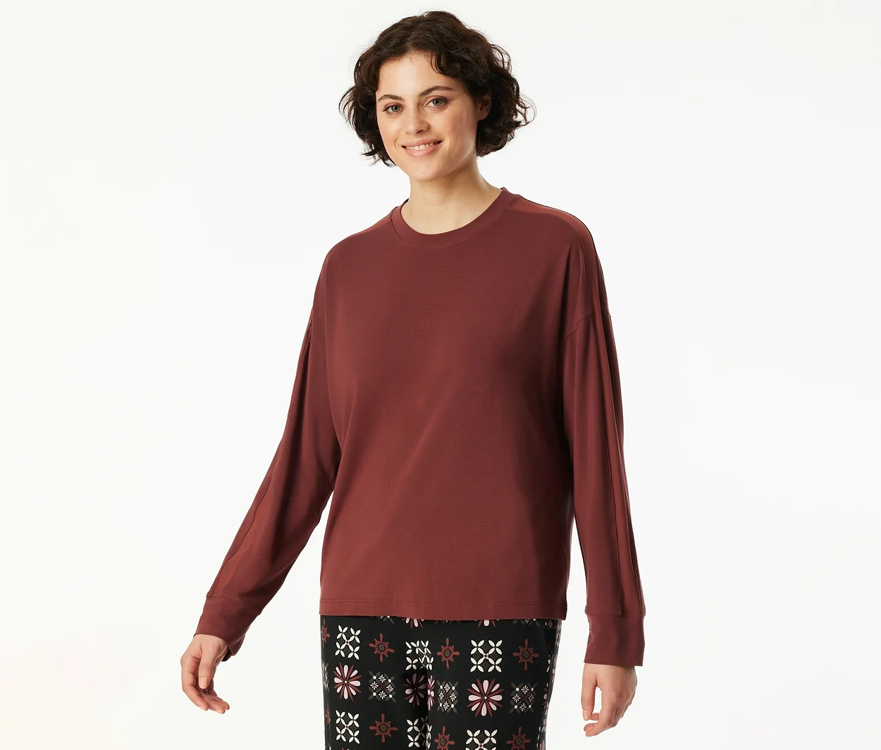 Eine lächelnde Frau mit kurzen, dunklen Haaren trägt ein weinrotes Langarmshirt und eine schwarze Hose mit Blumenmuster.