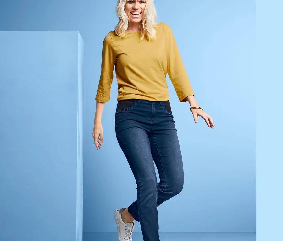Blonde Frau posiert in Jeans mit Komfortbund, gelbem Oberteil und weißen Schuhen vor blauem Hintergrund.
