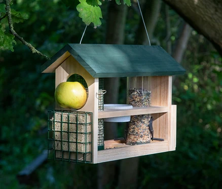 Ein hölzernes 7-in-1-Futterhaus hängt an einem Baum und ist mit Äpfeln, Talg und Vogelfutter gefüllt.