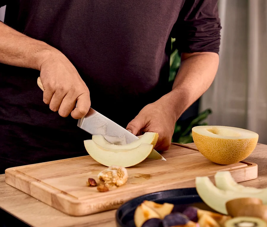 Person schneidet Melone mit Henckels by ZWILLING Pakka Messer auf Henckels Holzschneidebrett. Geschnittenes Obst auf Teller.