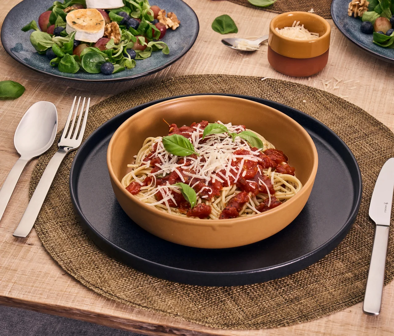Spaghetti mit Tomatensauce, Käse und Basilikum in einer Schüssel auf einem schwarzen Teller und Platzset. Das Henckels by ZWILLING Clouds Besteckset (Löffel, Gabel, Messer) liegt daneben. Salat im Hintergrund.