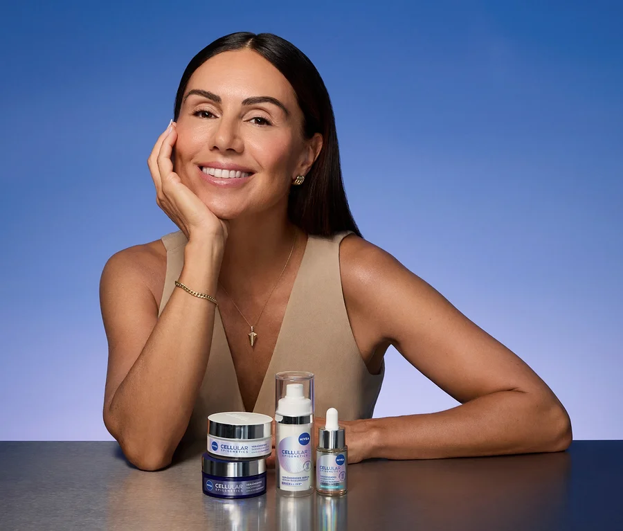 Eine lächelnde Frau mit dunklen Haaren, beigem Oberteil und Goldschmuck stützt ihr Kinn auf die Hand. Vor ihr stehen zwei Nivea Cellular Cremetiegel, eine Pumpflasche und eine Pipettenflasche auf einer reflektierenden Fläche.