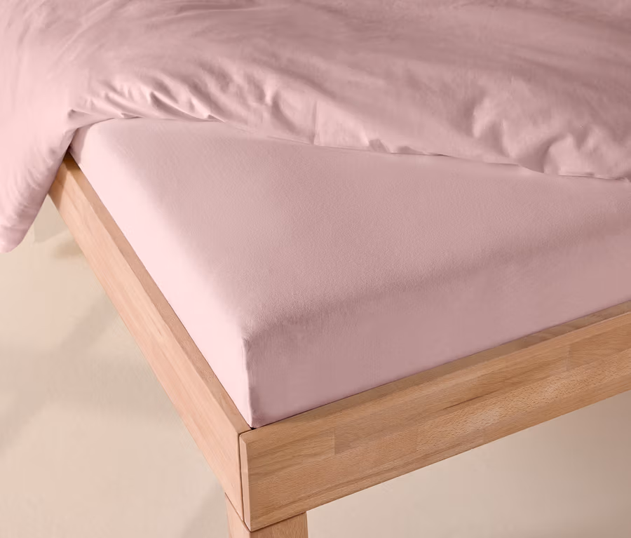 Rosafarbenes Jersey-Spannbettlaken auf einem Bett mit hellem Holzrahmen, teilweise von einer rosafarbenen Bettdecke bedeckt.