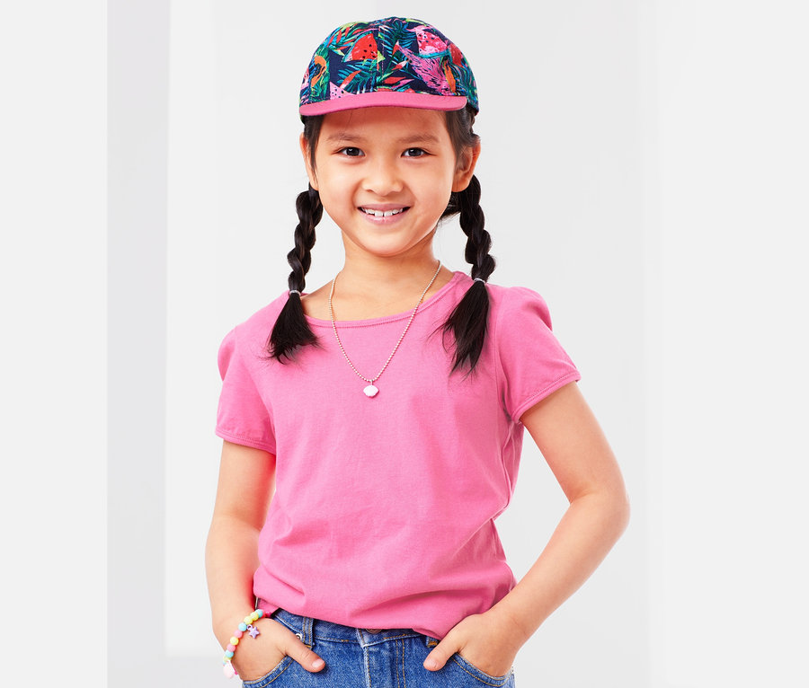 Lächelndes Mädchen mit Zöpfen, trägt ein rosa T-Shirt, einen Jeansrock und eine Cap.