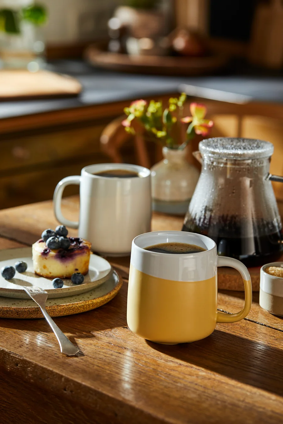 Auf einem Holztisch stehen eine Tasse Kaffee, ein Teller mit Kuchen und Blaubeeren und eine Glaskanne.