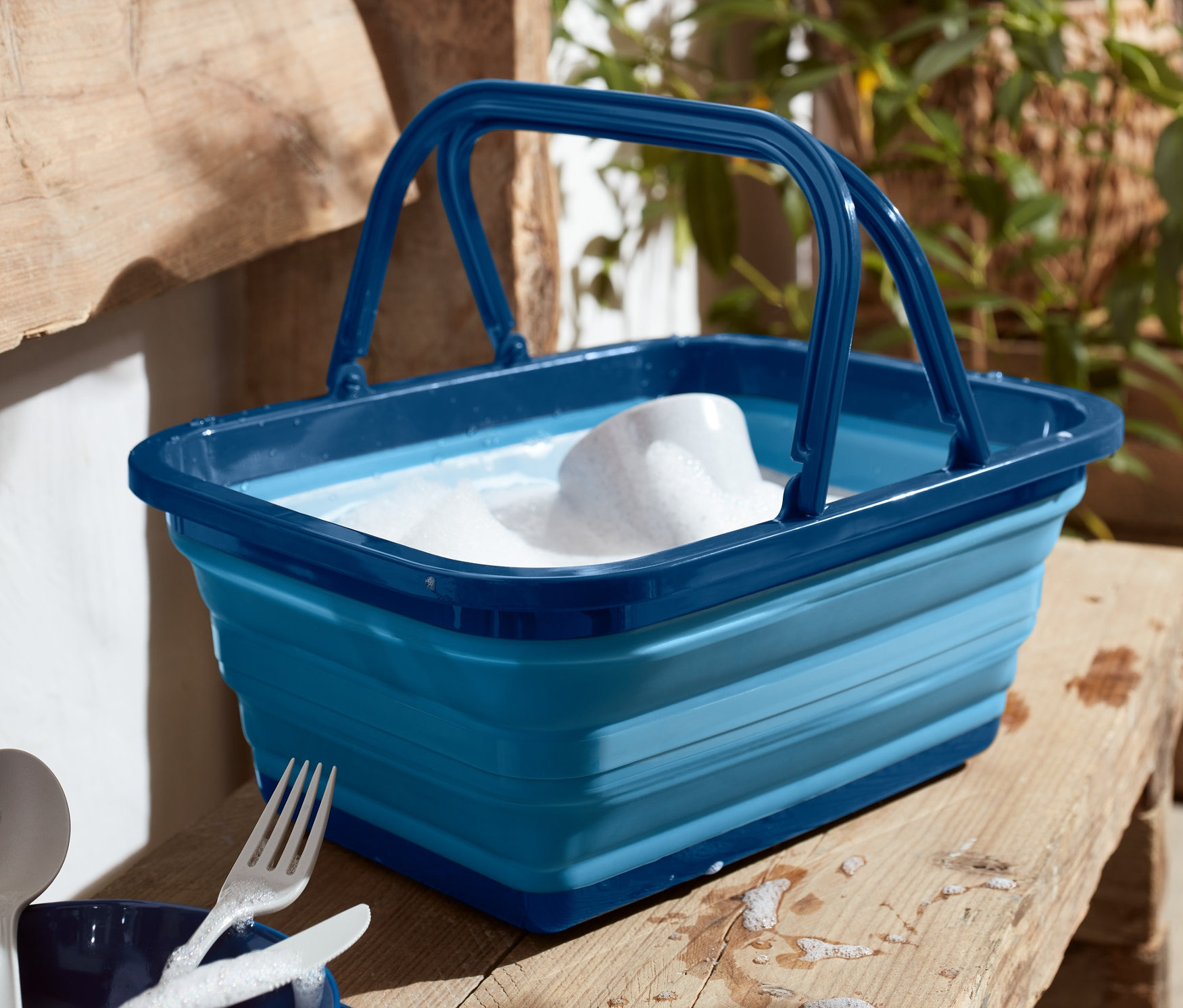 Eine blaue faltbare Spülschüssel mit Schaum auf einer Holzbank, daneben liegen blaue Teller und Besteck.