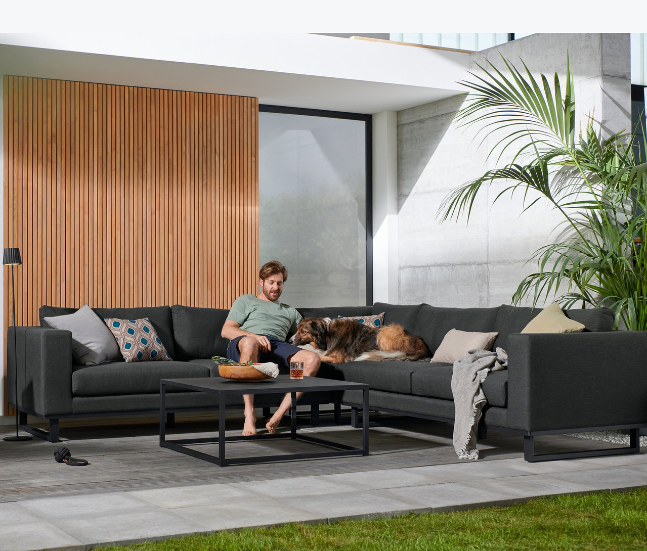Mann sitzt mit Hund auf anthrazitfarbener Premium-Lounge-Ecke mit Sunbrella®-Stoff. Eine LED-Akku-Stehleuchte und Outdoor Kissen, 4er-Set, grau stehen auf dem Tisch.