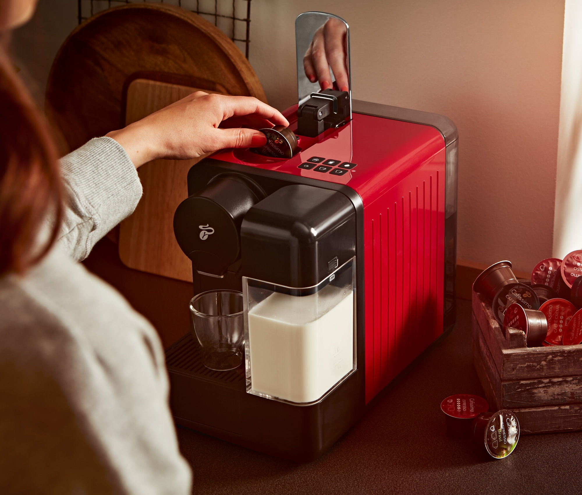 Frau legt Kapsel in rote Cafissimo milk Kaffeemaschine. Daneben stehen ein Glas Milch und eine Holzkiste mit Kapseln.