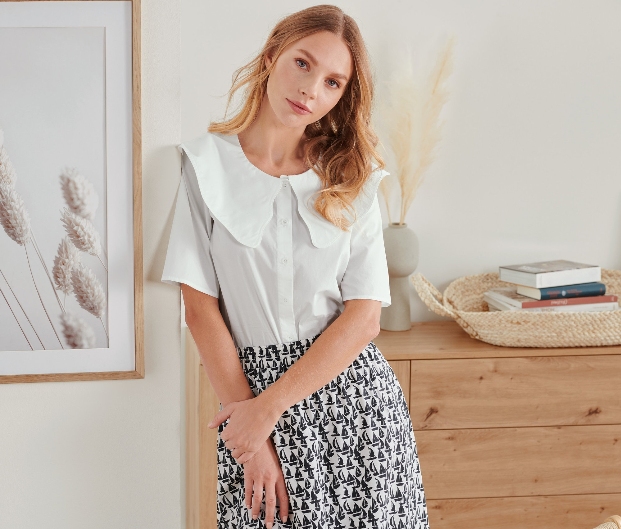Eine Frau lehnt an einer Wand und trägt eine Bluse mit Kragen und einen bedruckten Rock.