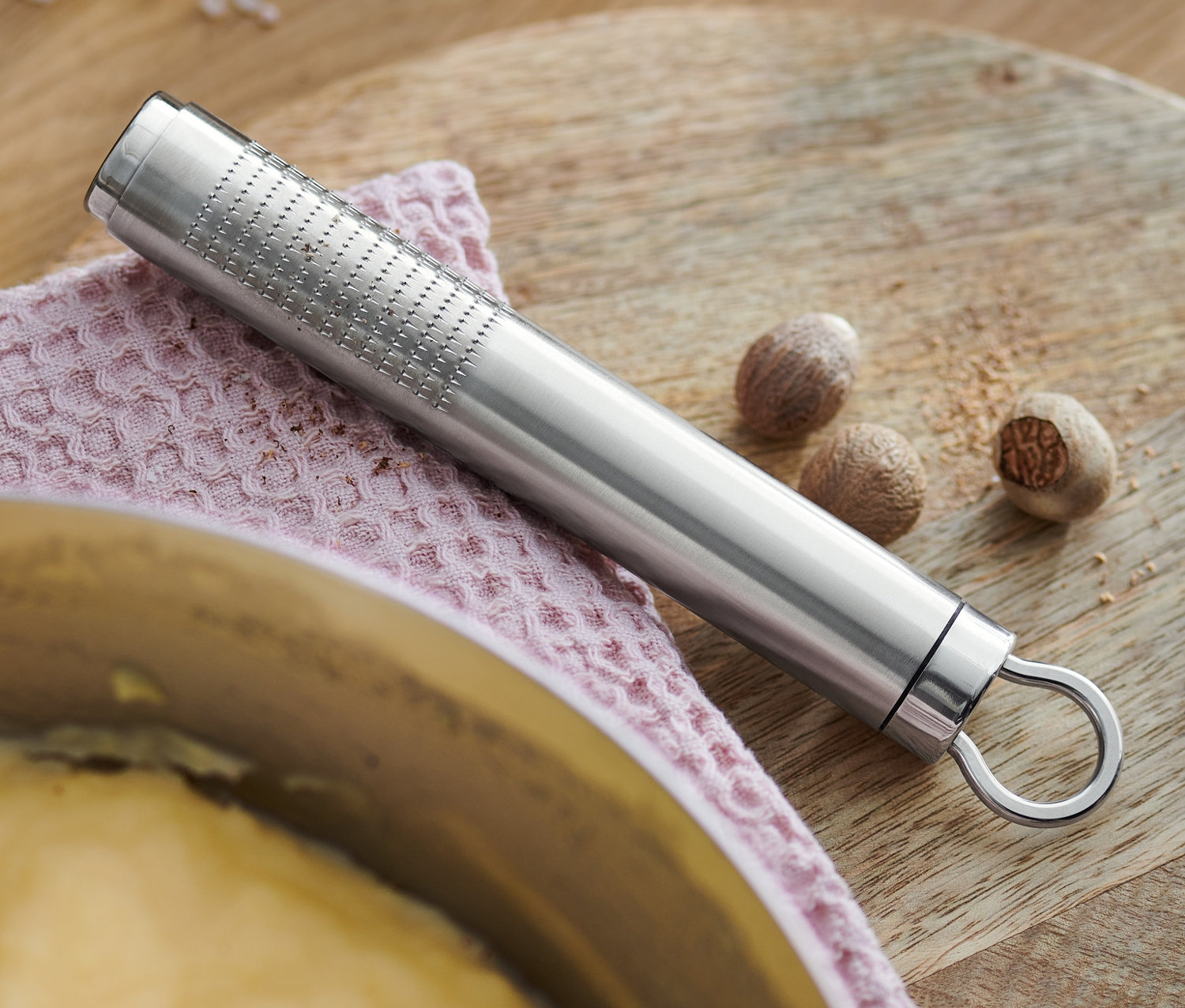 Eine Muskatreibe aus Edelstahl liegt auf einem rosa Tuch neben Muskatnüssen und einem Topf mit Brei.
