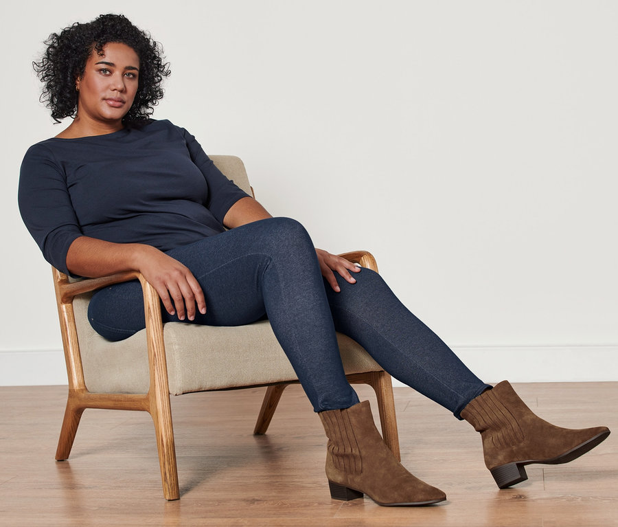 Frau sitzt im Sessel und trägt ein dunkelblaues Shirt mit 3/4-Arm, eine gefütterte Leggings in Jeansoptik und Stiefeletten aus Veloursleder.