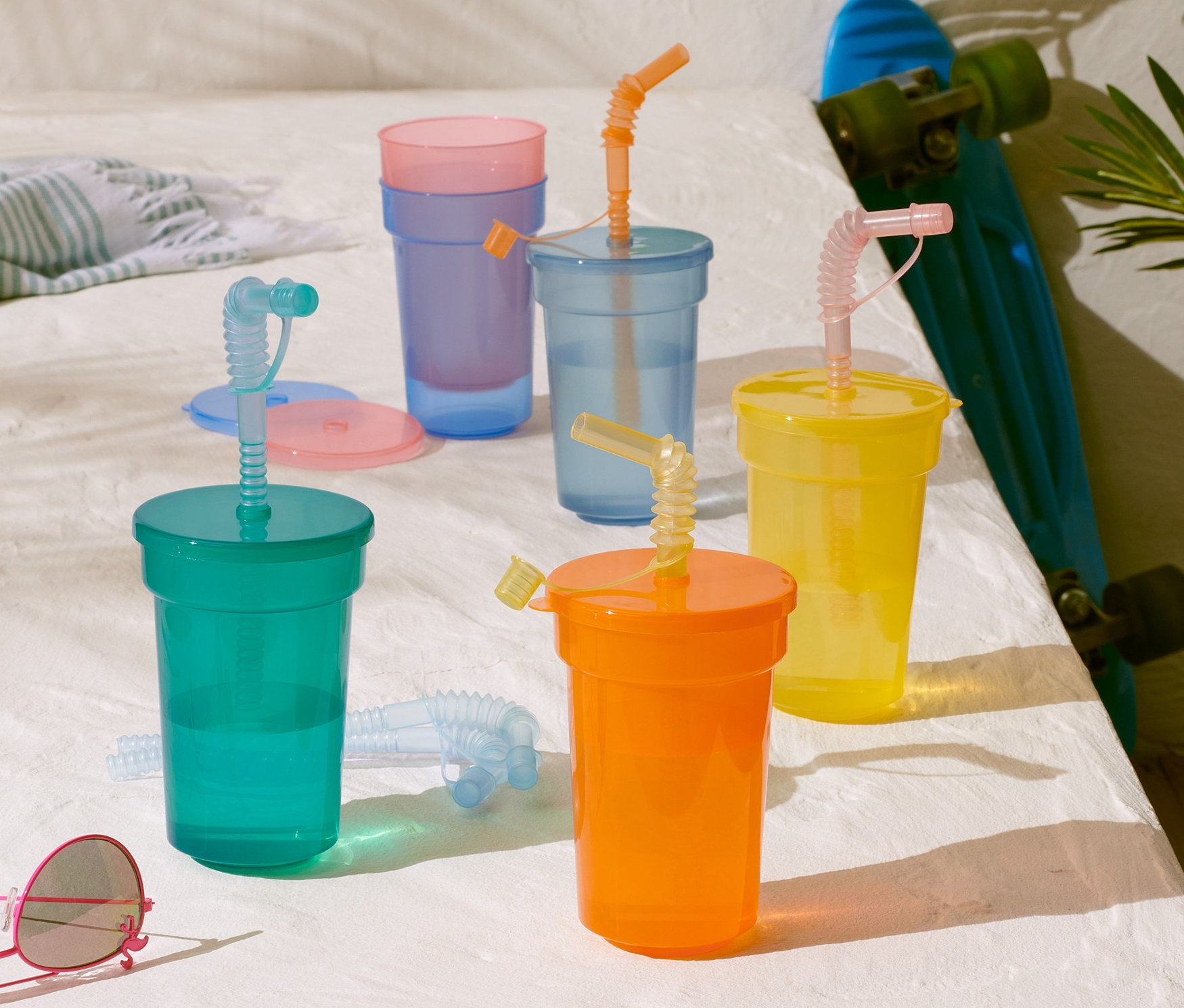 Ein Kinderbecher-Set mit Strohhalmen in verschiedenen Farben auf einem weißen Tisch neben einem Skateboard und einer Sonnenbrille.