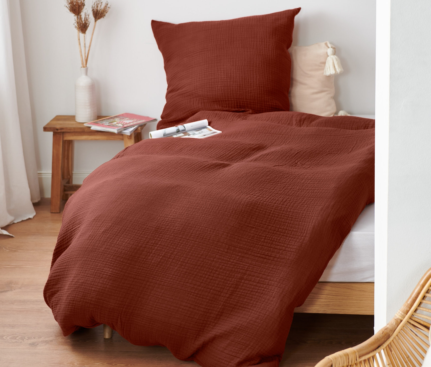 Rotbraune Musselin-Bettwäsche Normalgröße auf einem Bett mit Holzrahmen. Auf dem Nachttisch steht eine Vase mit Blumen.