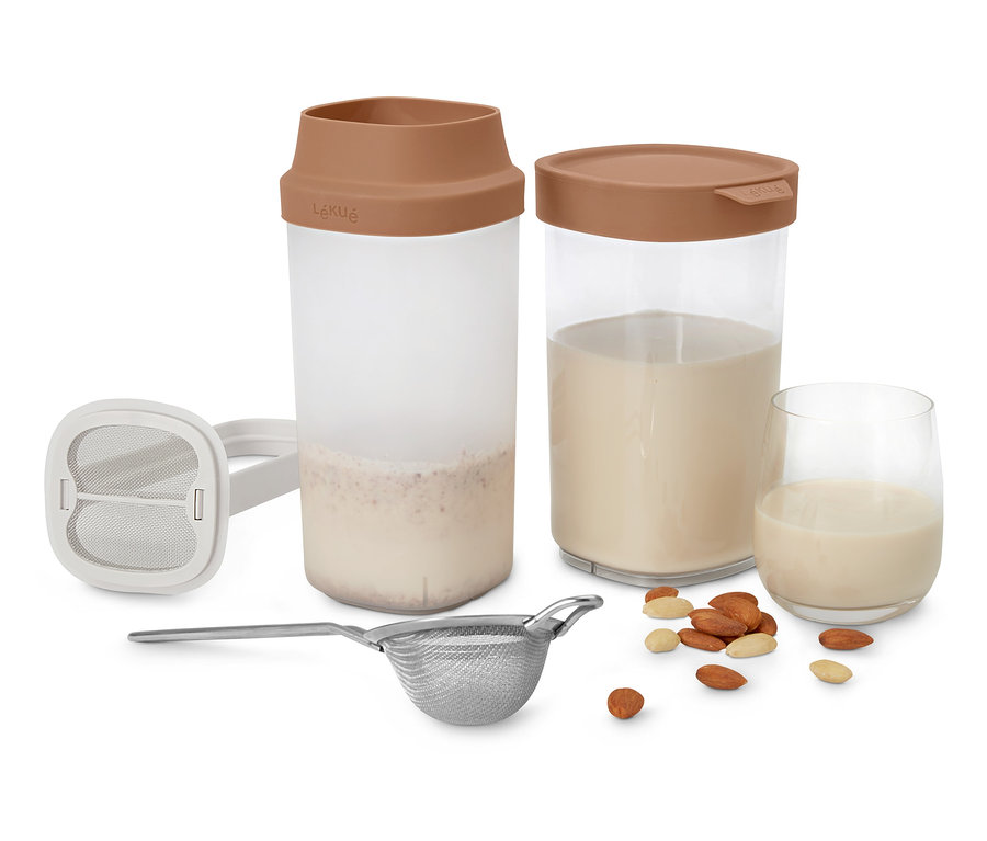 Zwei Lékué Nuss-und-Getreide-Milch-Maker, Sieb und ein Glas Nussmilch mit Mandeln auf weißem Hintergrund.