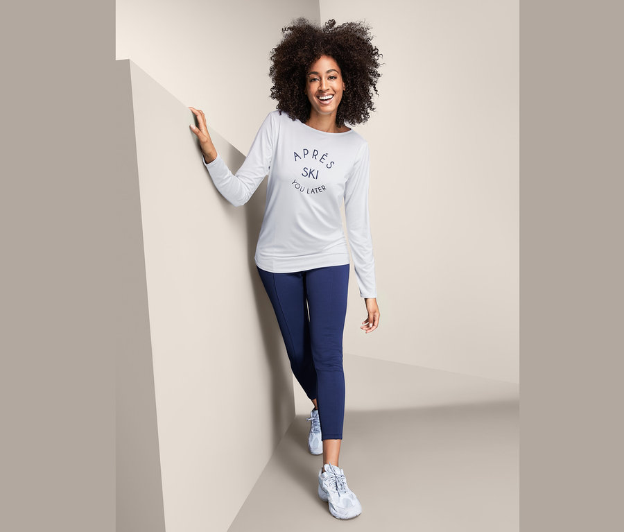Eine Frau mit lockigem Haar lächelt und posiert in einem weißen Funktionsshirt mit langen Ärmeln und dunkelblauen Leggings.