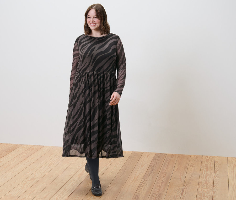 Frau geht über einen Holzboden und trägt ein bedrucktes Mesh-Kleid und 2 Paar Feinstrumpfhosen, 80 den, schwarz.