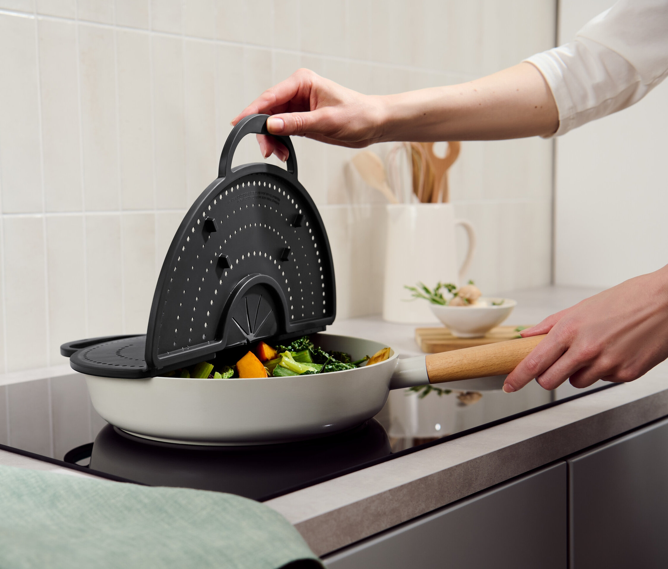Hand hält Pfannenspritzschutz über einer Pfanne mit Gemüse.