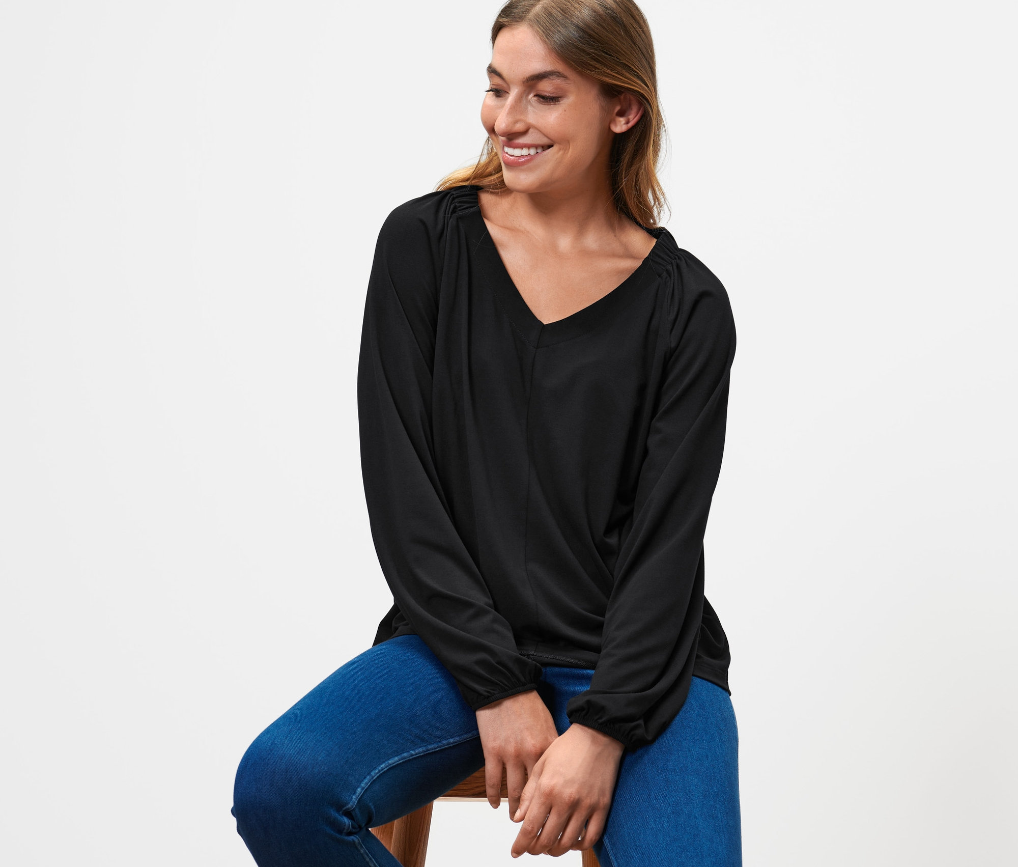 Frau sitzt auf einem Hocker und trägt ein schwarzes Blusenshirt mit Raffung und blaue Jeans.