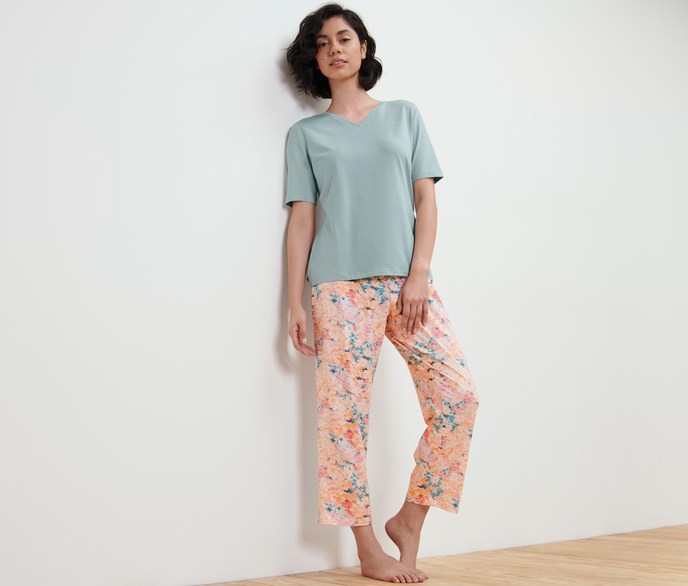 Eine Frau lehnt an einer Wand und trägt einen Pyjama, eine Hose mit Blumen-Alloverprint und ein hellgrünes T-Shirt.