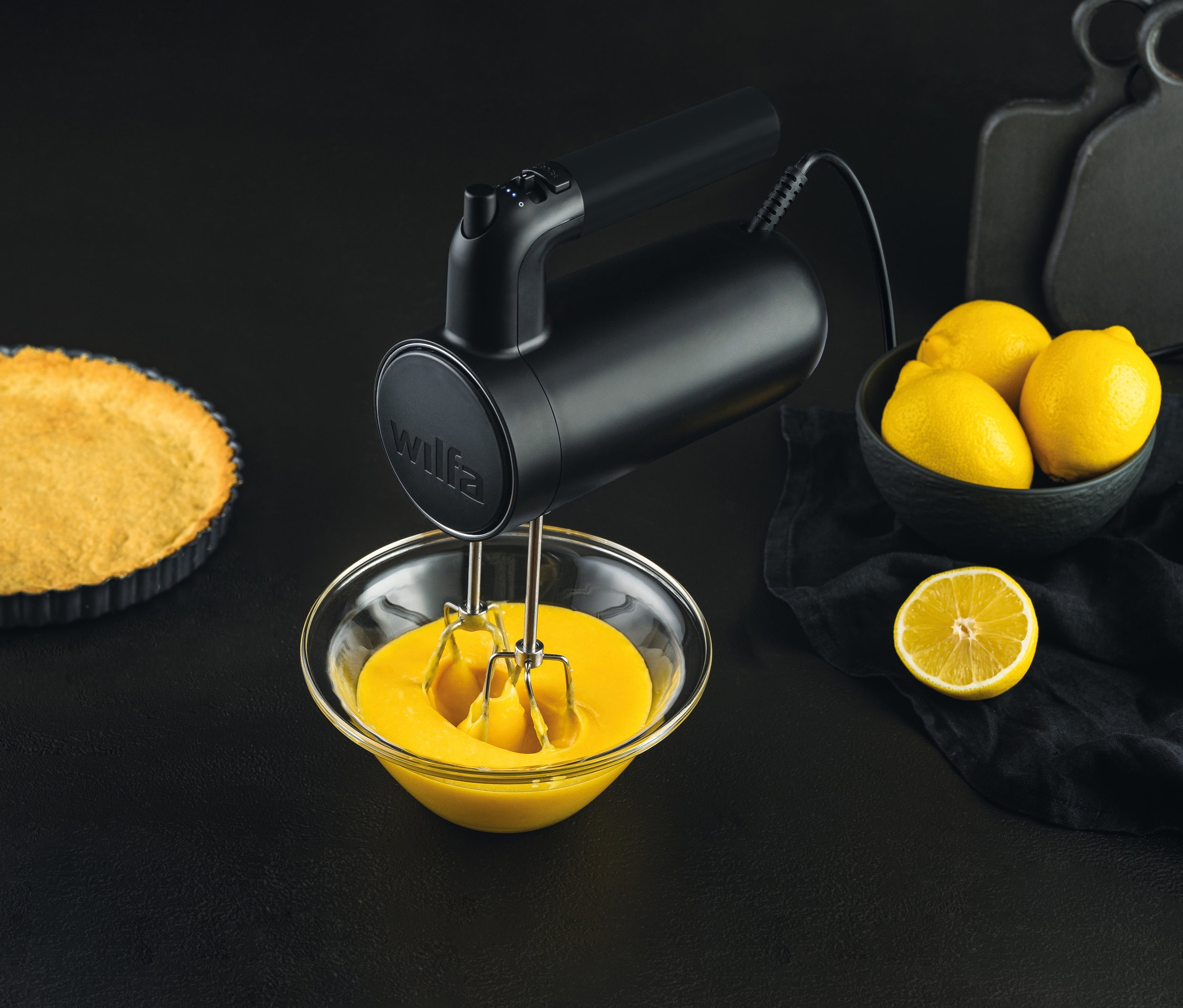 Schwarzer WILFA Handmixer SMOOTH-MIX »HM1B-50« mixt gelbe Masse in Schüssel neben gebackenem Kuchenboden und Schale mit Zitronen.
